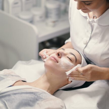 Woman lying during cosmetic procedure while skillful female cosmetologist making facial treatment